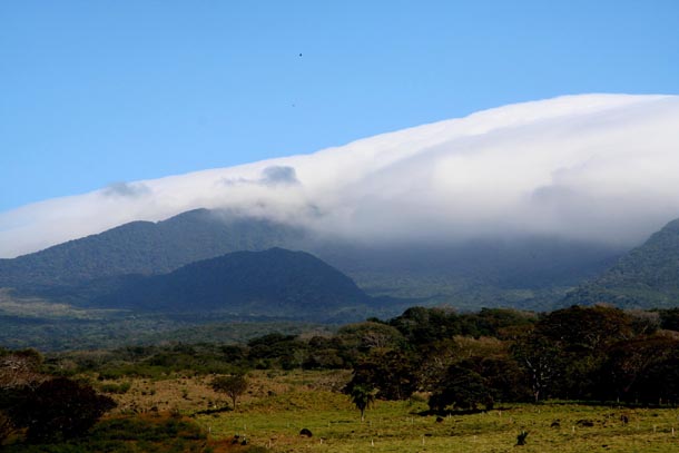 Side view of a blown-out volcano caldera, with forest on the steep slopes and thick clouds on the top