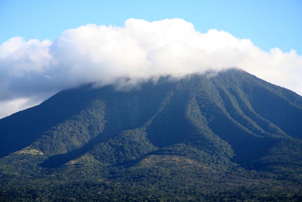A single forested volcano, Volcan Orosi, with steep rugged slopes, topped with a white cloud that is large and dark gray to the left (east) and thin and fluffy white to the right (west)