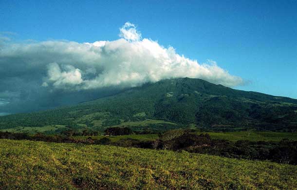 A sprawling forested volcano, Rincon, from afar topped with clouds in a blue sky. On the left (east side) the clouds are thick and dark, on the right they vanish into wisps in a blue sky